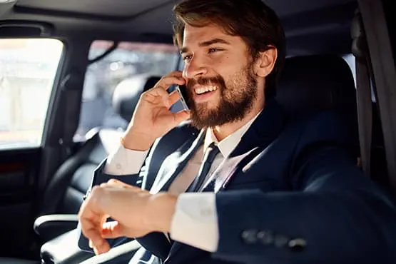Chauffeur en costume, souriant au téléphone depuis l’habitacle d’une berline.